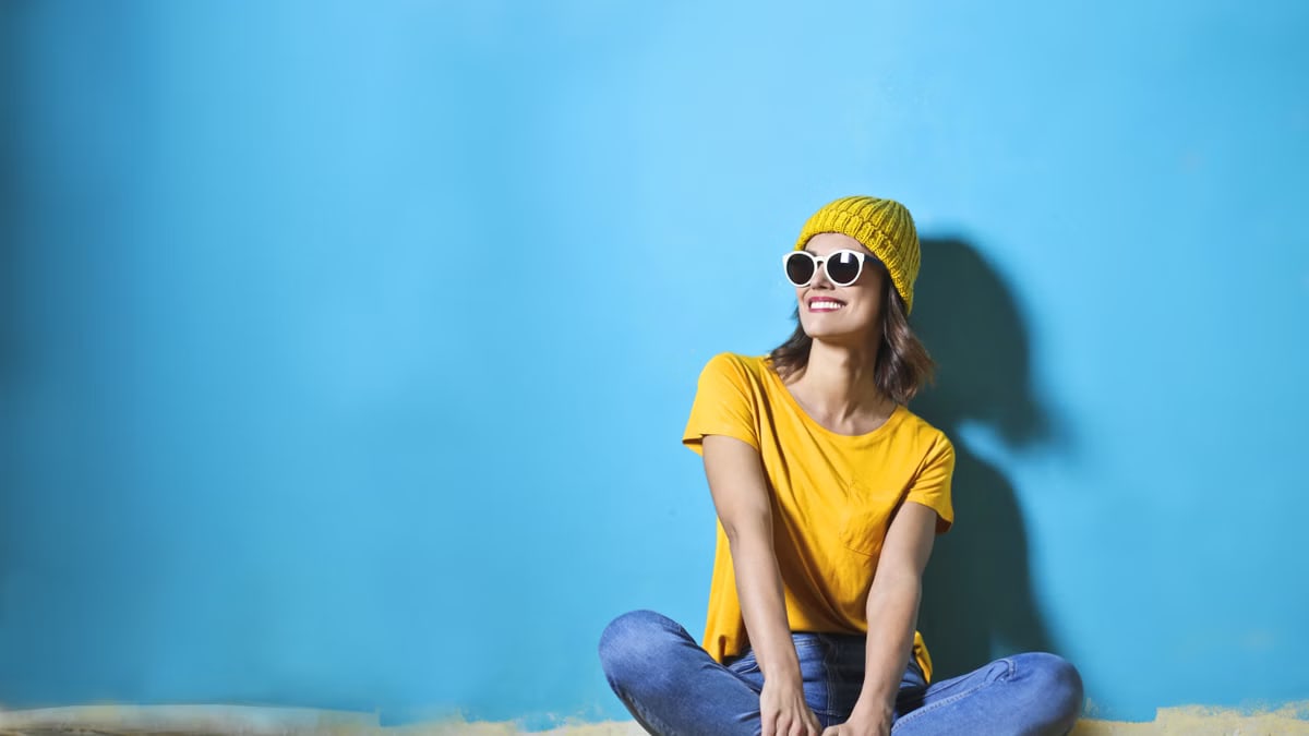 Fashionable young woman sitting indoors wearing sunglasses and a yellow top on a bright blue background.